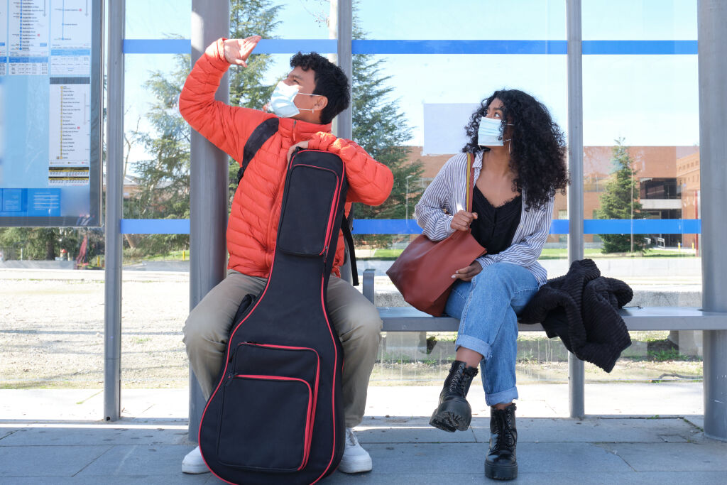 Young latin couple wearing protective face mask and carrying guitar case looking at the bus, sitting at bus stop. New normal in public transport.